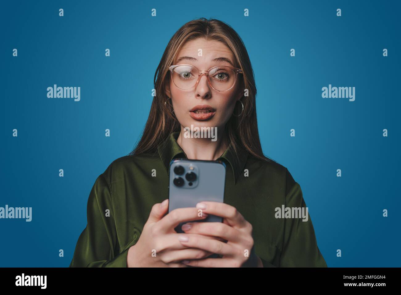 Une femme choquée qui réagit à un sms frustrant sur un téléphone portable, se plaignant du texte d'un smartphone, debout sur fond bleu. Banque D'Images
