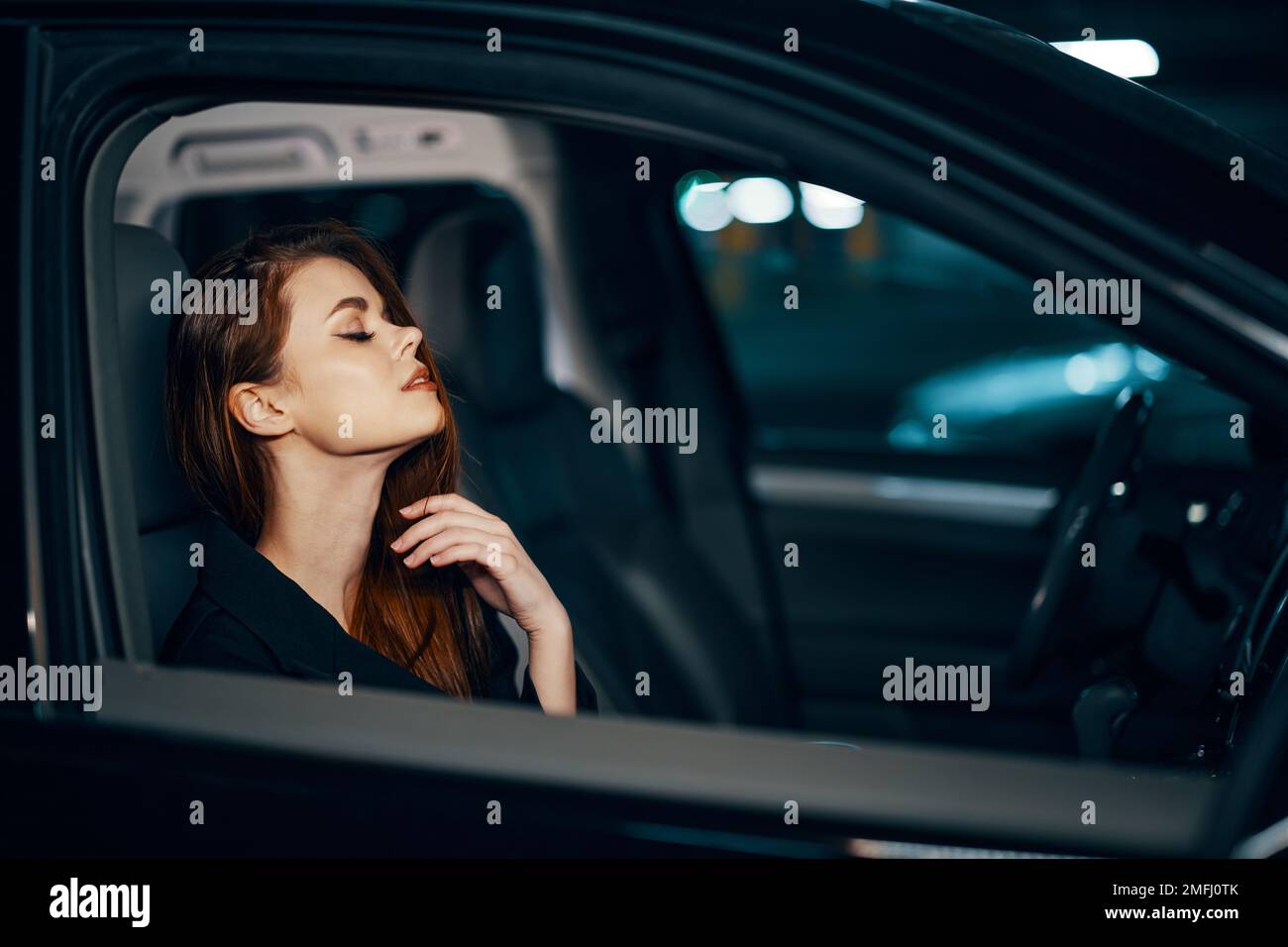 une femme élégante et luxueuse est assise dans une voiture noire la nuit, redressant ses cheveux longs et stylisés, fermant ses yeux avec plaisir Banque D'Images