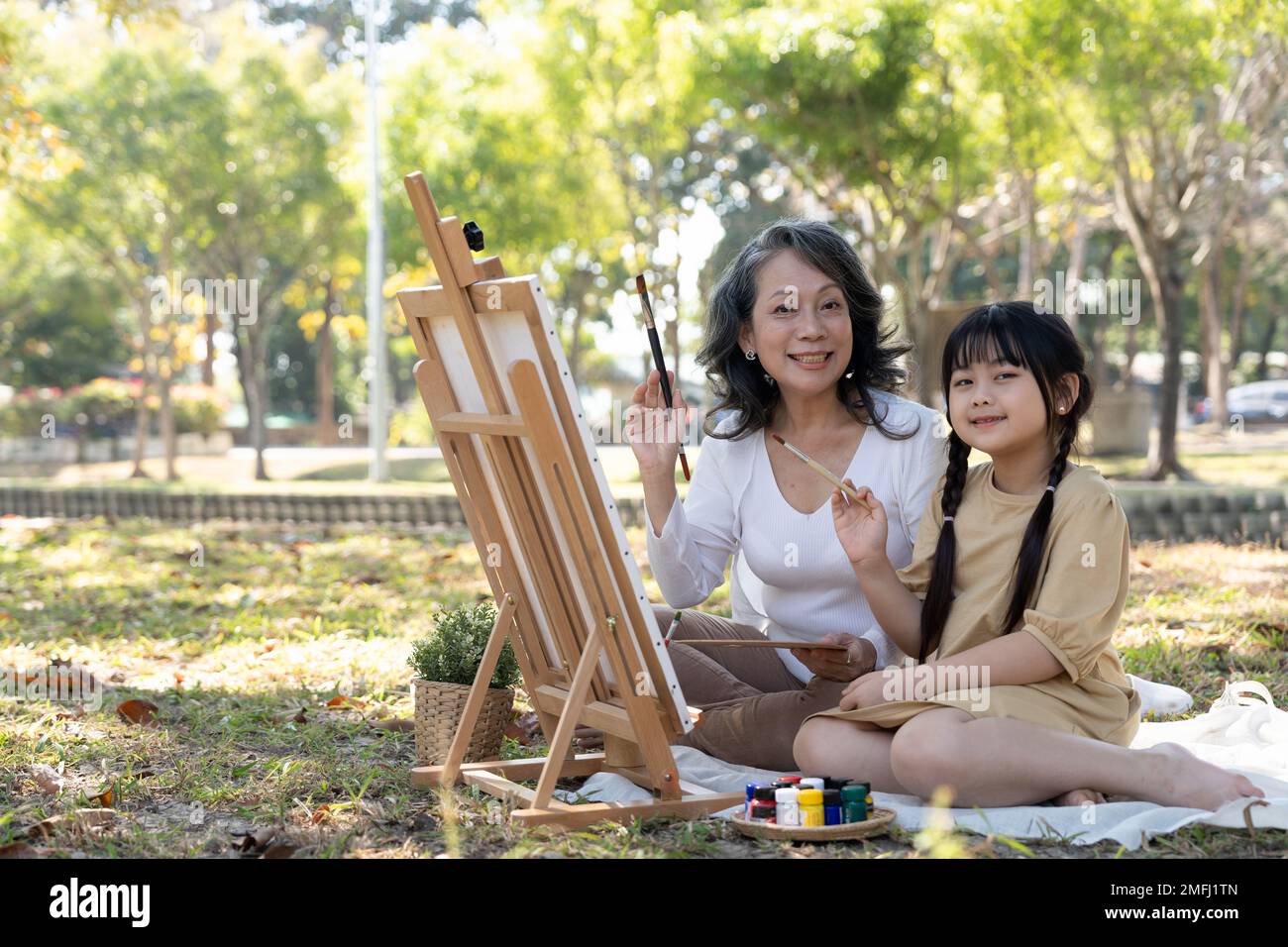 Une belle et heureuse grand-mère asiatique enseignant à sa petite-fille de peindre des aquarelles sur toile, ayant un grand moment ensemble dans le beau parc Banque D'Images