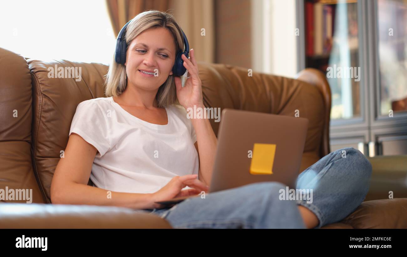 Femme avec un casque souriant et regardant l'écran d'un ordinateur portable tout en étant assise sur le canapé. Banque D'Images