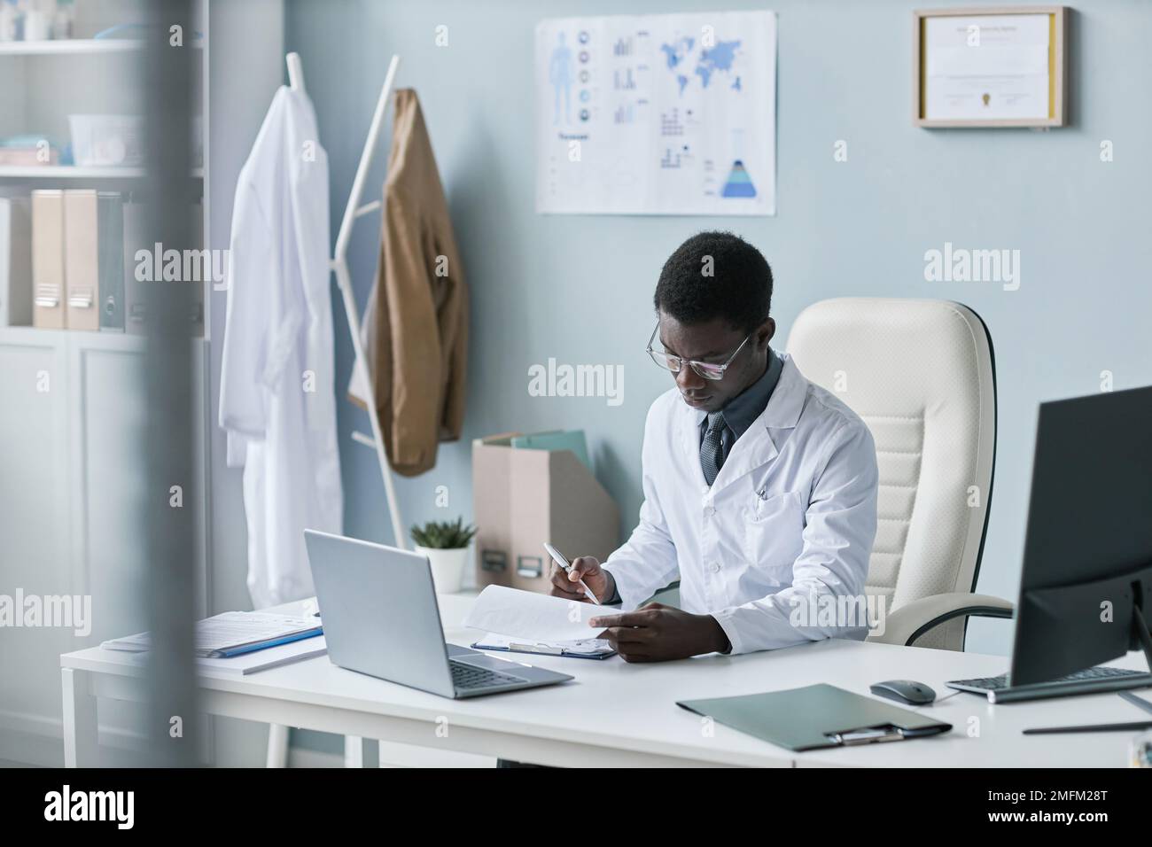 Portrait d'un jeune médecin noir travaillant avec des documents au bureau et portant un blouse de laboratoire Banque D'Images