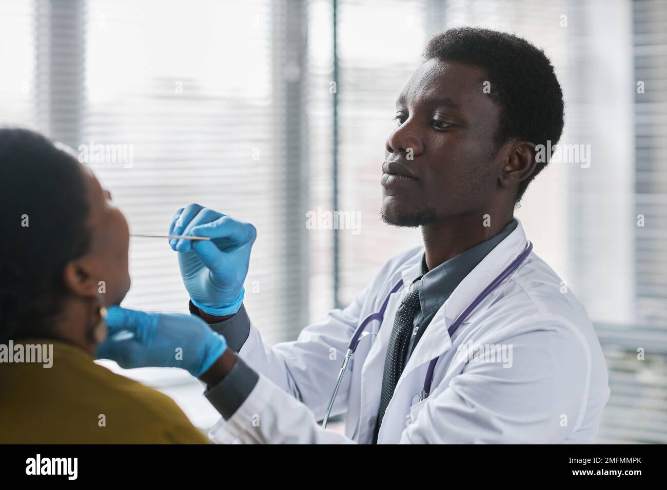 Portrait de la vue latérale d'un jeune médecin noir consultant un patient en clinique et effectuant un examen de la gorge pour la grippe Banque D'Images
