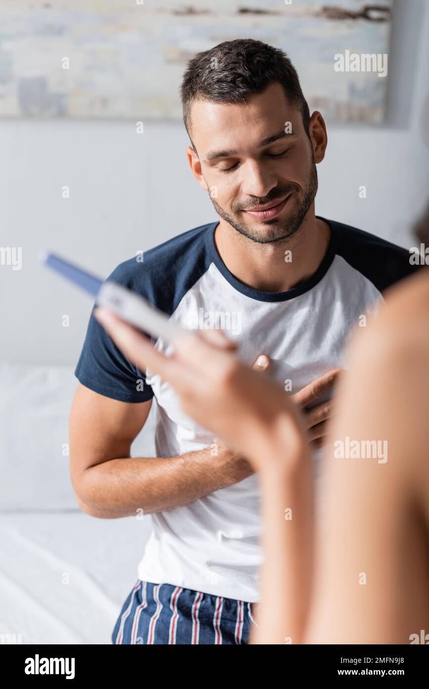 Homme souriant dans un pyjama debout près d'une petite amie avec test de grossesse dans la chambre, image de stock Banque D'Images