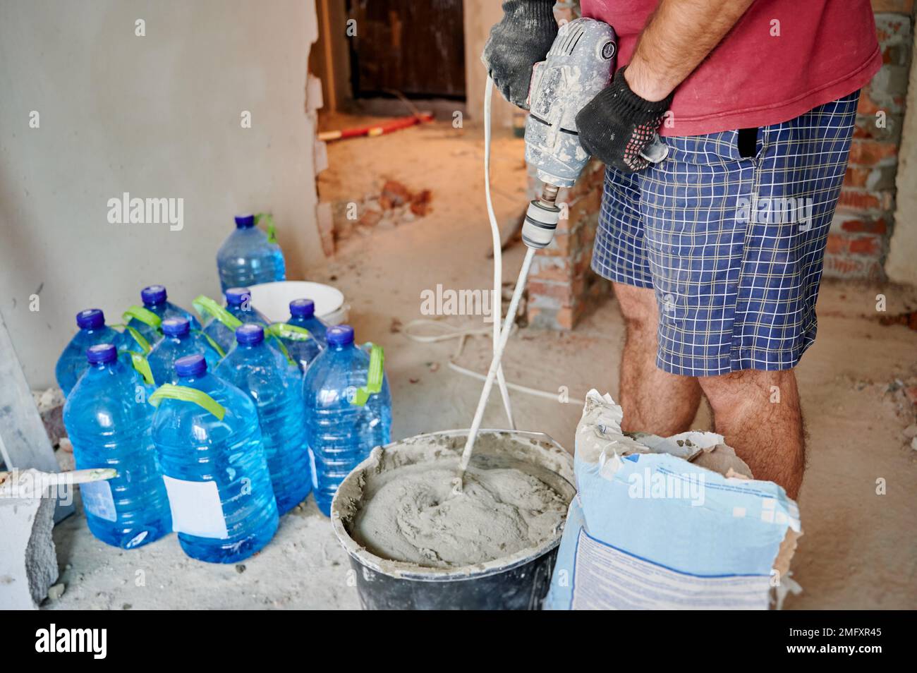 Gros plan de l'homme constructeur dans les gants de travail de préparation du mélange sable-ciment pour briqueter dans le bâtiment en construction. Travailleur masculin mélangeant du mortier avec un mélangeur électrique de ciment pour béton. Banque D'Images
