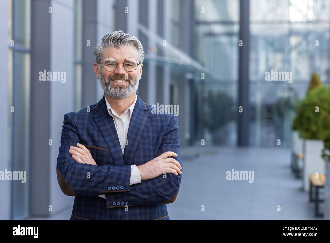 Portrait d'un homme d'affaires mûr aux cheveux gris, d'un patron senior souriant et regardant un appareil photo aux bras croisés, d'un homme aux cheveux gris à l'extérieur du bâtiment de bureau, d'un investisseur et d'un propriétaire prospères en costume d'affaires. Banque D'Images