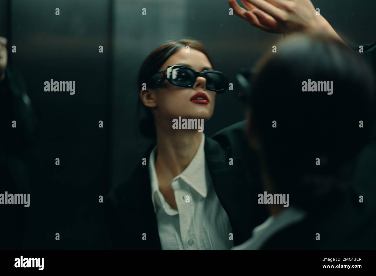 Femme élégante dans une veste noire et des lunettes de soleil se posant dans un ascenseur près du miroir, modèle de mode, lumière cinématique sombre et matrice de couleurs Banque D'Images