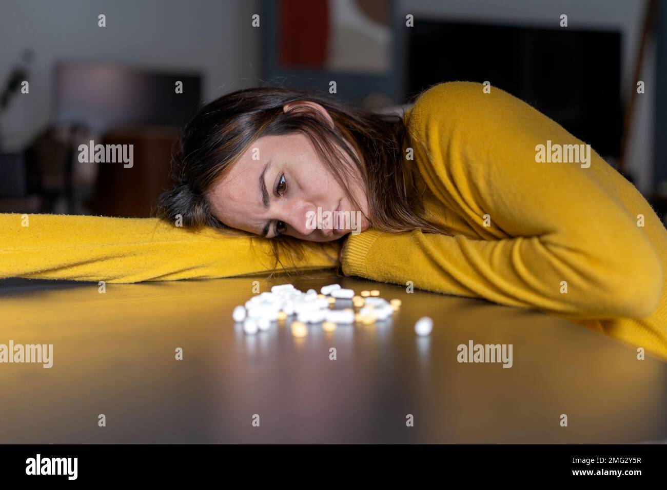 Femme mélancolique adulte avec de longs cheveux foncés dans un chandail jaune, tête penchée à la main à la table avec pile de pilules antidépresseurs à la maison Banque D'Images