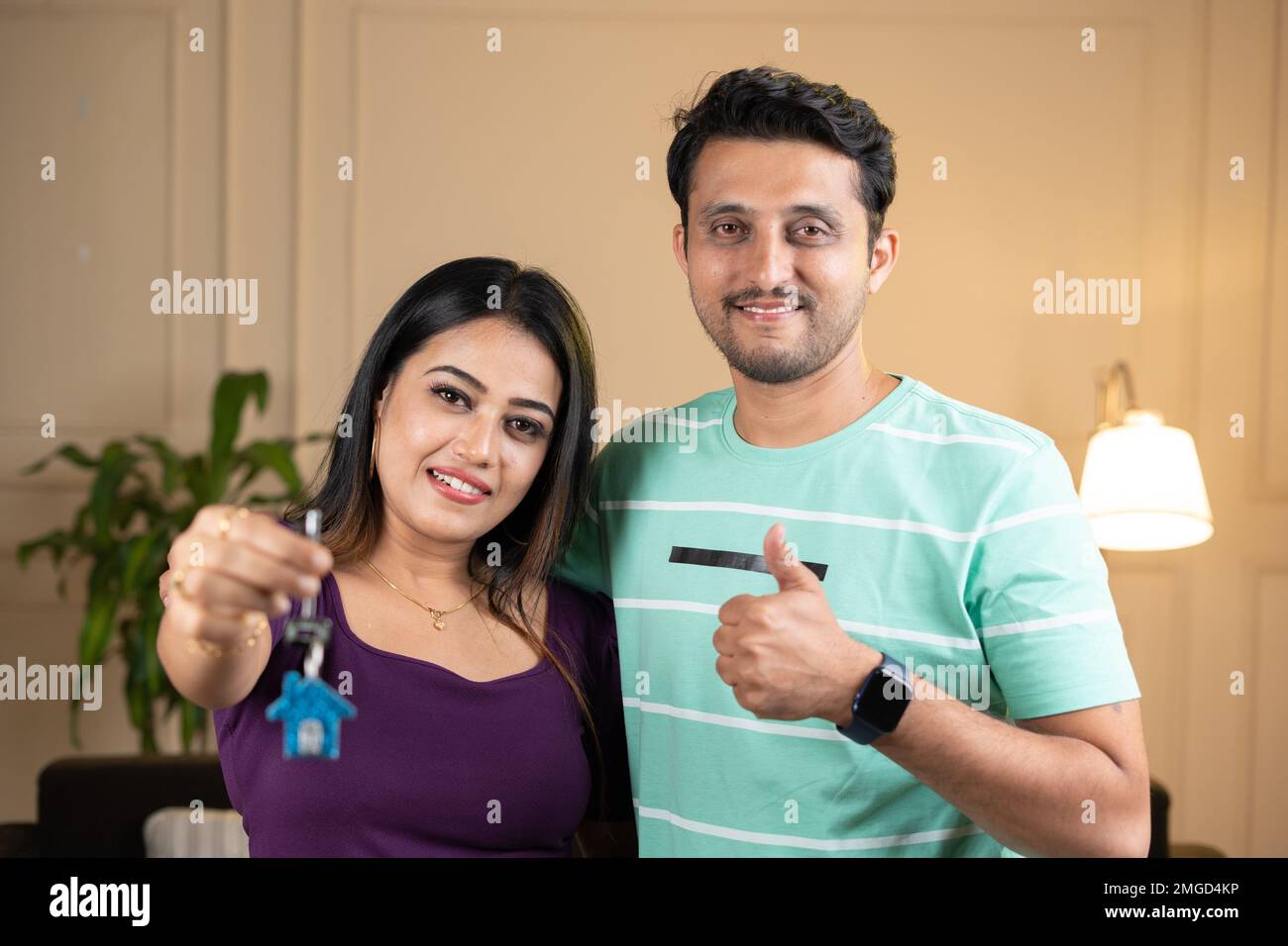 Femme souriante avec husbnd montrant les clés de la maison avec le pouce vers le haut geste en regardant l'appareil photo à la maison - concept d'assurance, l'achat de la nouvelle maison et Banque D'Images
