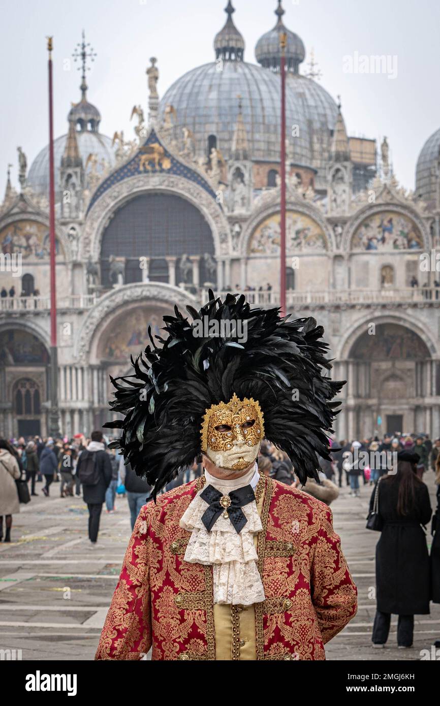Un homme d'âge moyen en costume rouge vintage avec col en dentelle, portant un masque de carnaval bordé d'un cercle de plumes noires élevées, se tient seul au Th Banque D'Images