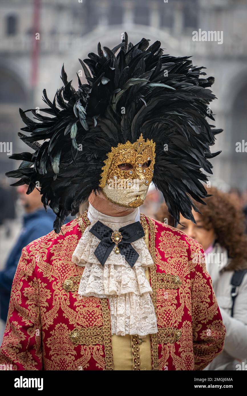 Un homme d'âge moyen en costume rouge vintage avec col en dentelle, portant un masque de carnaval bordé d'un cercle de plumes noires élevées, se tient seul Banque D'Images