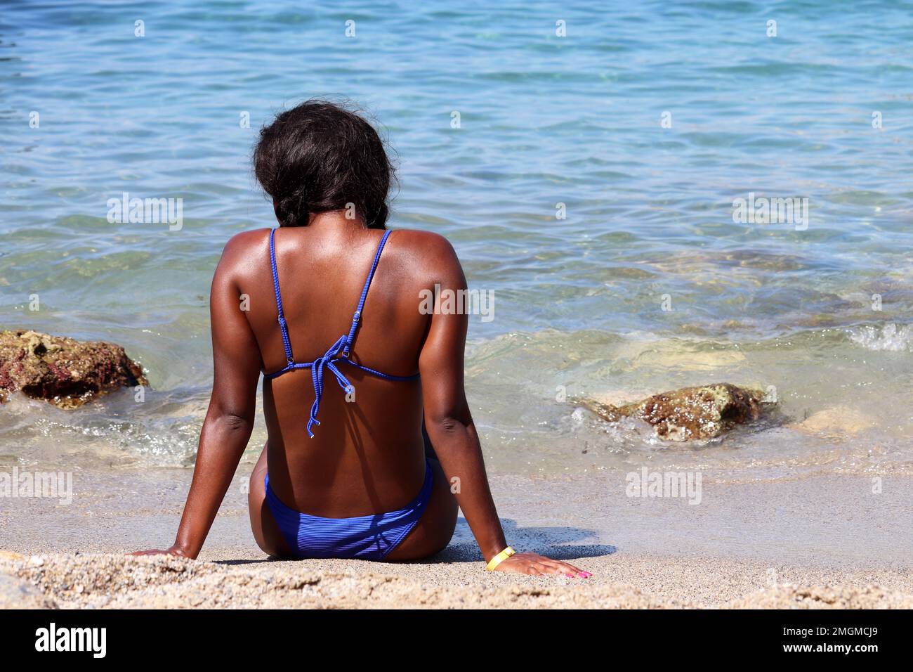 Vacances à la plage, femme noire en bikini bleu assis sur la côte de mer, vue arrière Banque D'Images