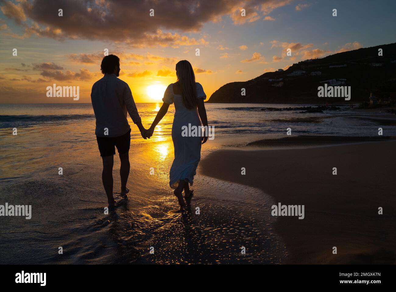 Jeune romantique habillé en couple marchant sur la plage de l'île vers le coucher de soleil pittoresque Banque D'Images