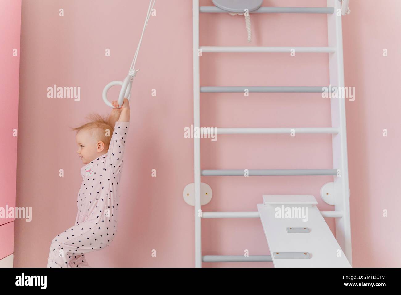 Une petite fille en pyjama rose doux fait de la gymnastique aux barres murales de la maison Banque D'Images