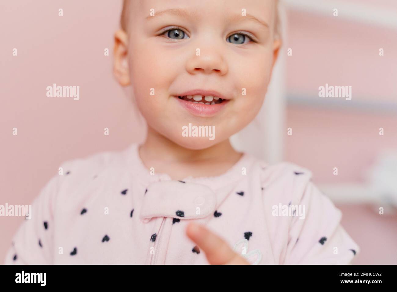 Portrait en gros plan d'une petite fille de race blanche en pyjama rose clair dans une chambre rose. Yeux gris, sourire avec les dents. Banque D'Images
