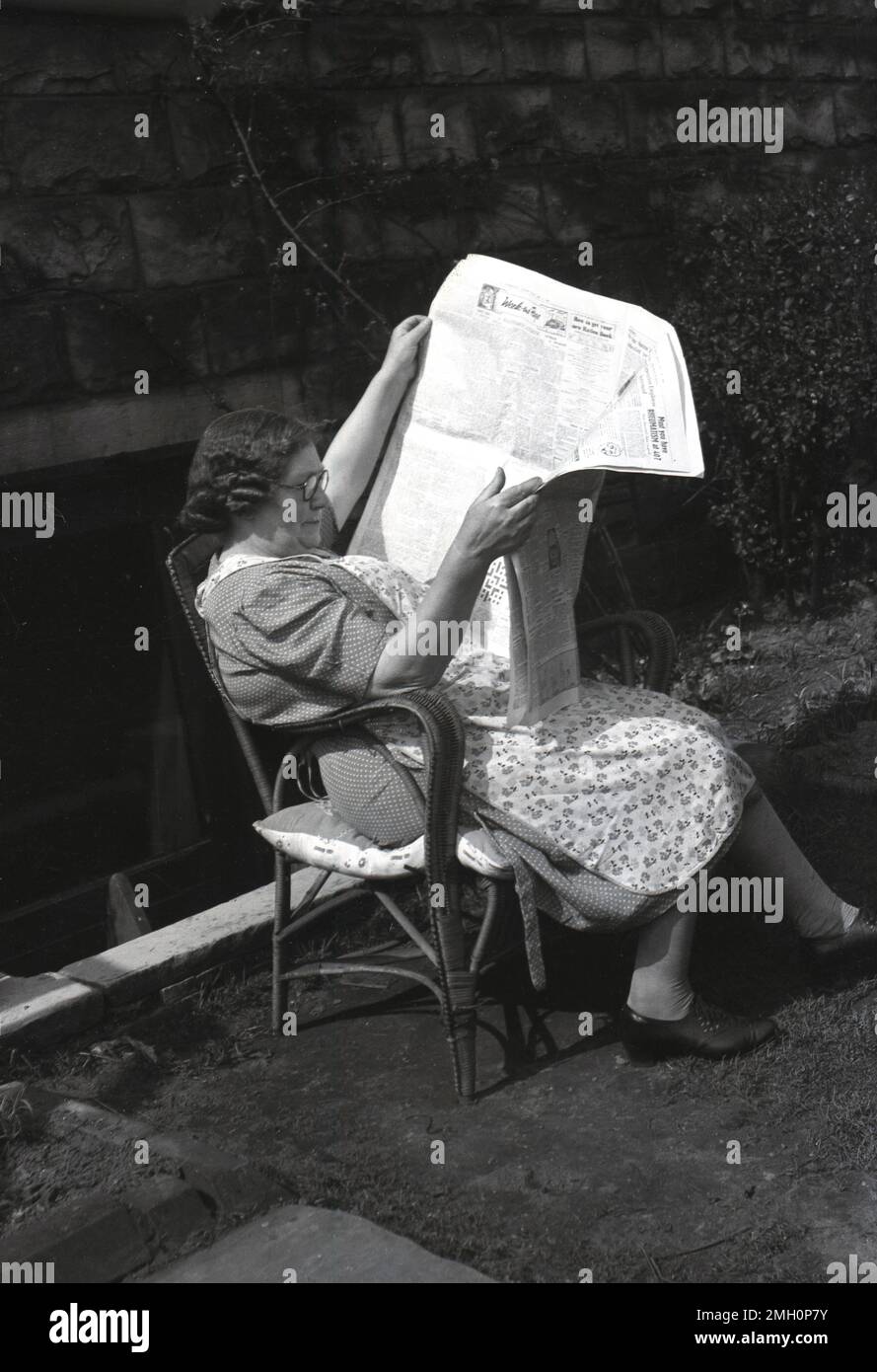 1950s, historique, par temps ensoleillé, une dame portant une robe à pois, avec un robe chasuble, assise sur une chaise en osier devant un grand journal, Angleterre, Royaume-Uni. Un tablier sans manches était courant à cette époque, notamment lors des travaux ménagers. Dans le journal, un titre dit, "Comment obtenir votre livre de nouvelle ration". Banque D'Images