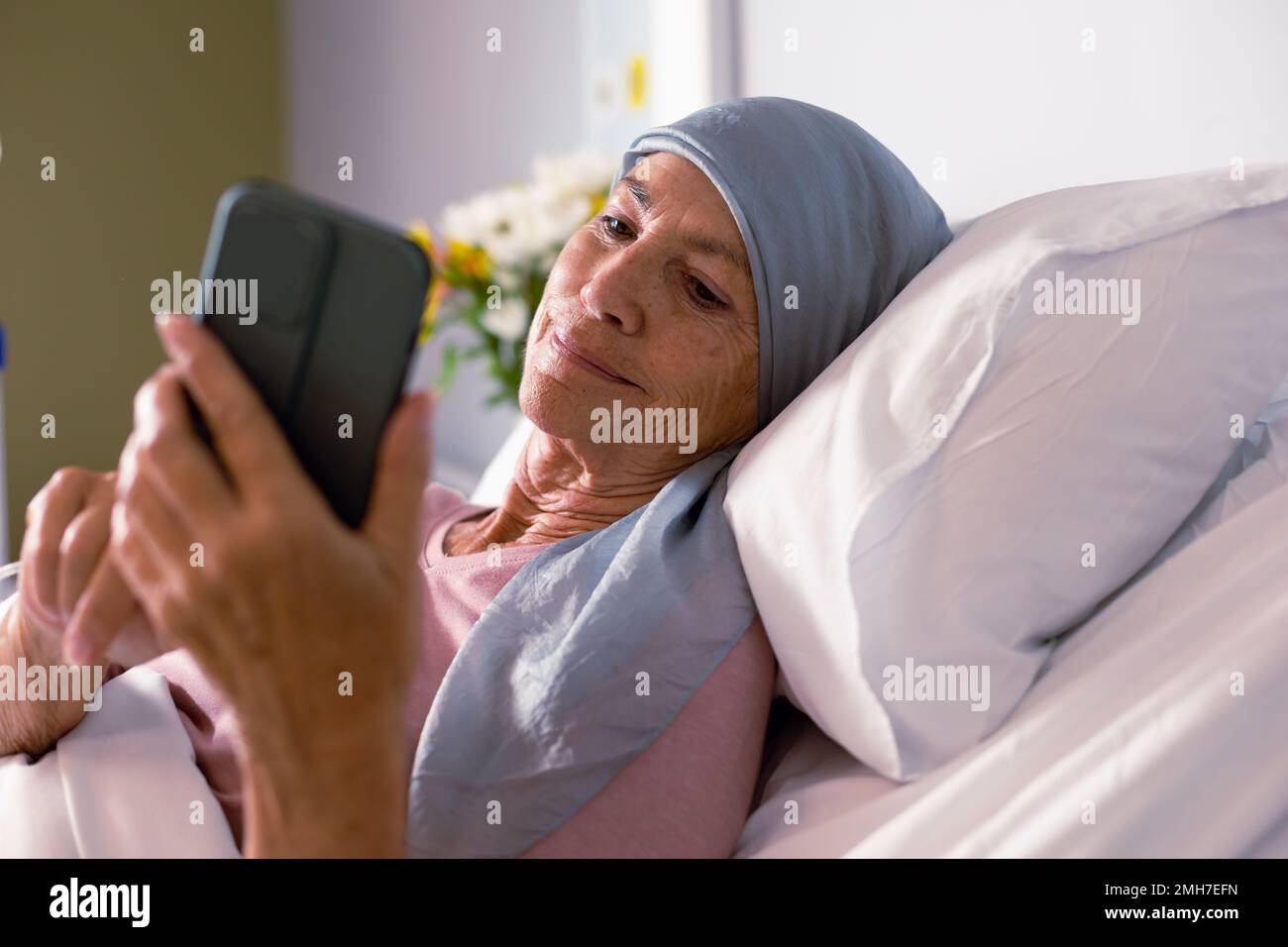 Patiente caucasienne âgée atteinte d'un cancer dans un foulard dans un lit d'hôpital à l'aide d'un smartphone Banque D'Images
