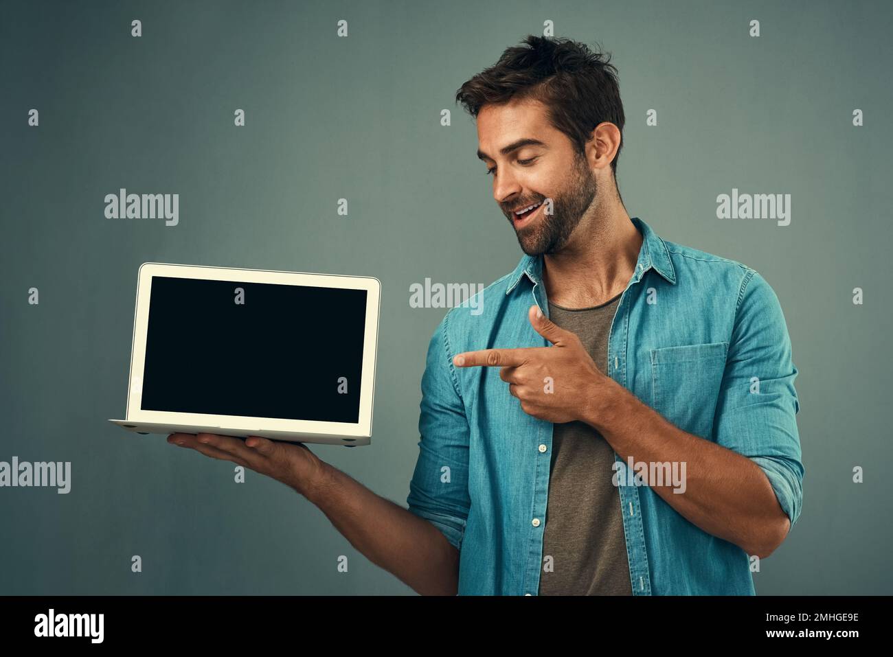 Découvrez-le ici. Studio photo d'un beau jeune homme pointant vers un ordinateur portable avec un écran vierge sur fond gris. Banque D'Images