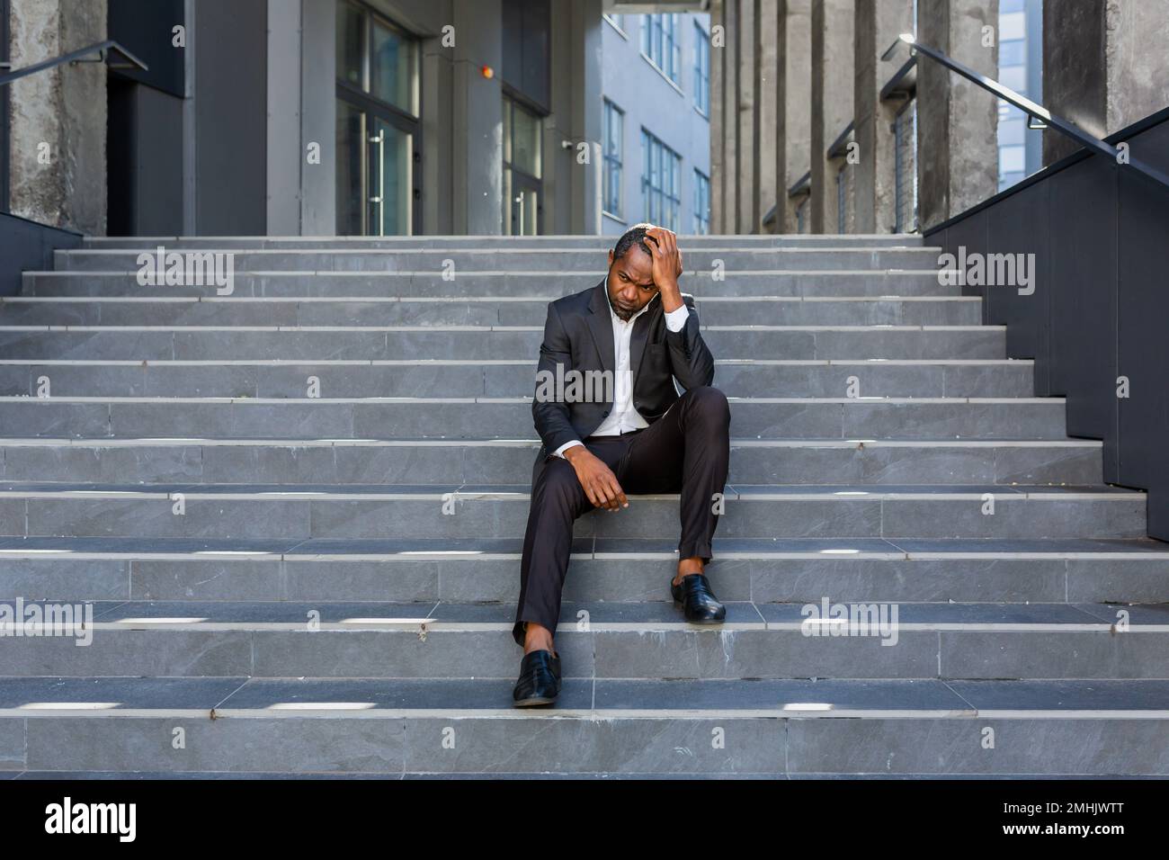 Contrarié afro-américain homme assis sur les escaliers à l'extérieur de l'immeuble de bureaux, homme d'affaires en faillite dans le désespoir, patron mature déprimé dans un costume d'affaires à l'extérieur. Banque D'Images