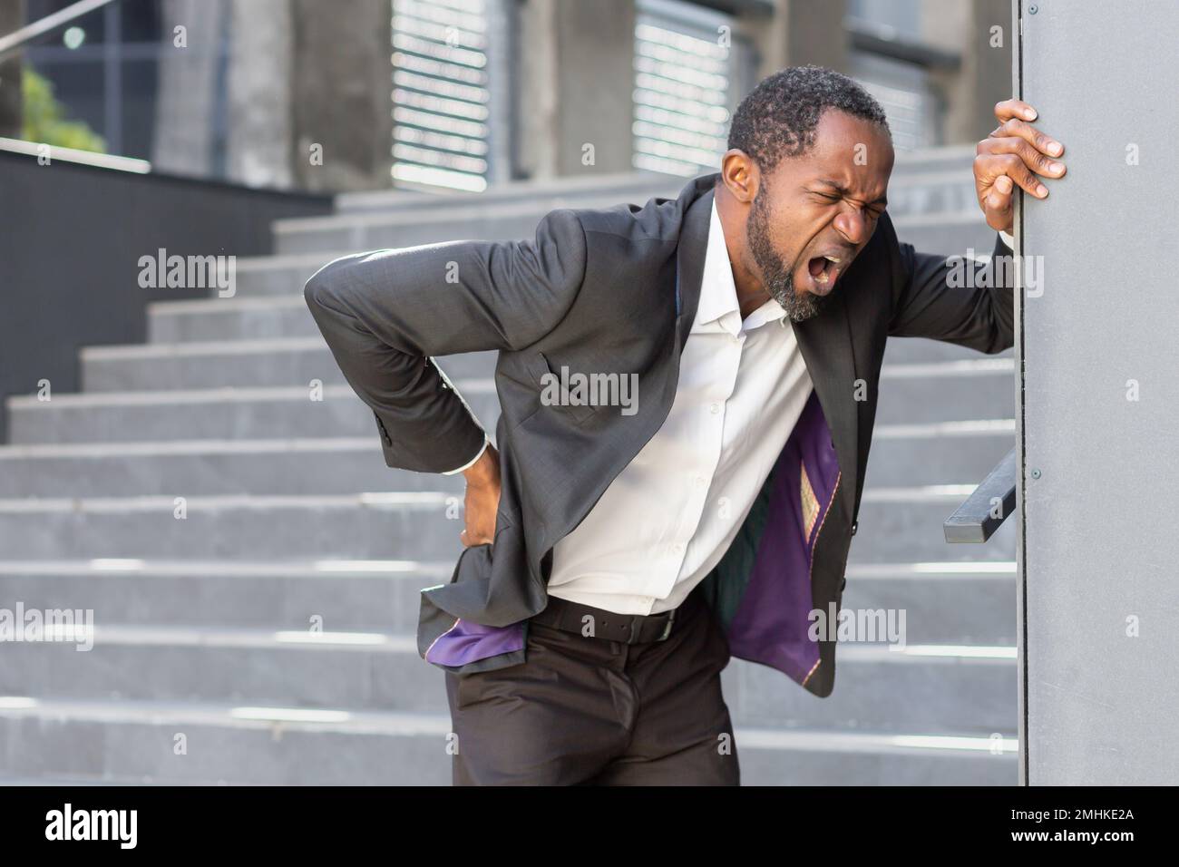 L'homme afro-américain surchargé et malade à l'extérieur du bâtiment de bureau a de graves maux de dos, homme d'affaires massant son côté, patron mature dans les airs près du bureau. Banque D'Images