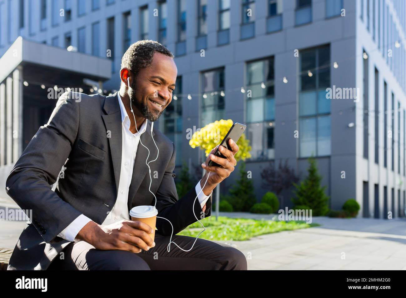 Homme d'affaires afro-américain utilisant le téléphone assis sur un banc à l'extérieur du bâtiment de bureau, le patron regardant la vidéo et souriant dans des écouteurs, homme à l'extérieur écoutant de la musique livres audio et des podcasts radio en ligne. Banque D'Images