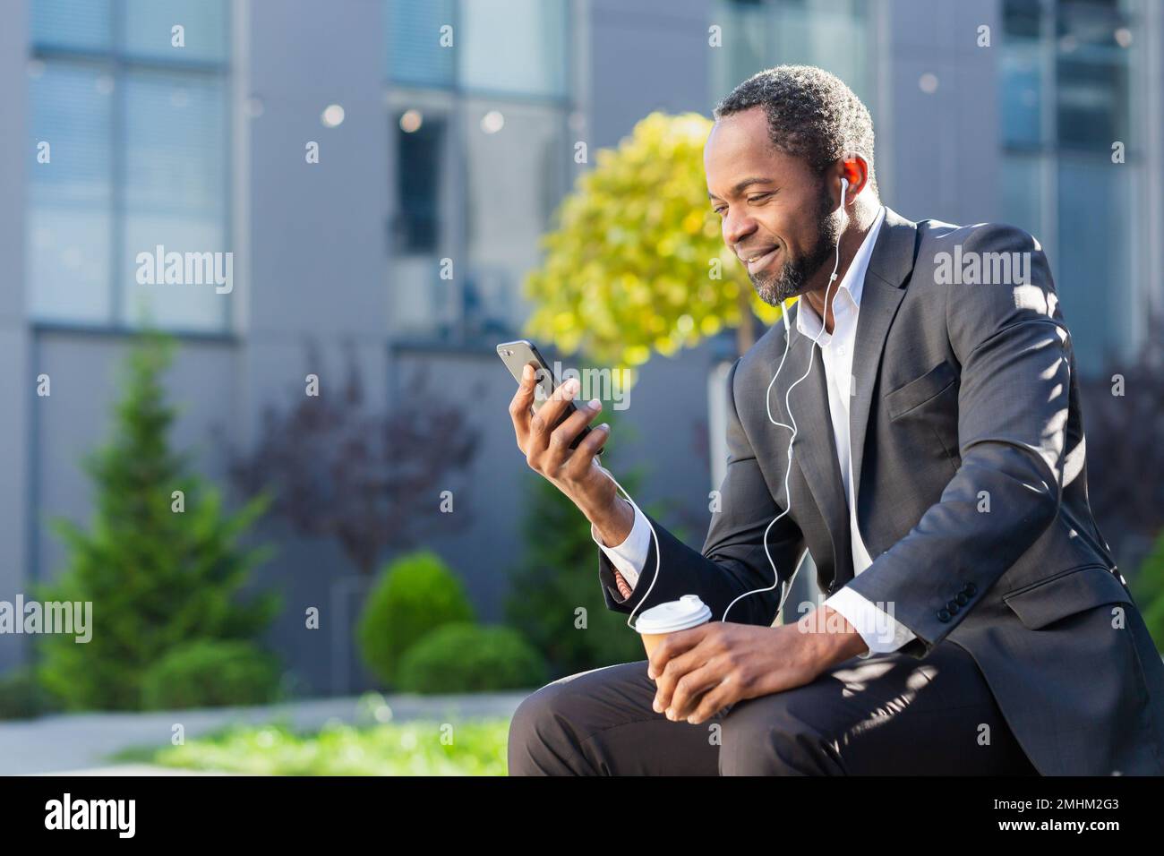 Homme d'affaires afro-américain utilisant le téléphone assis sur un banc à l'extérieur du bâtiment de bureau, le patron regardant la vidéo et souriant dans des écouteurs, homme à l'extérieur écoutant de la musique livres audio et des podcasts radio en ligne. Banque D'Images