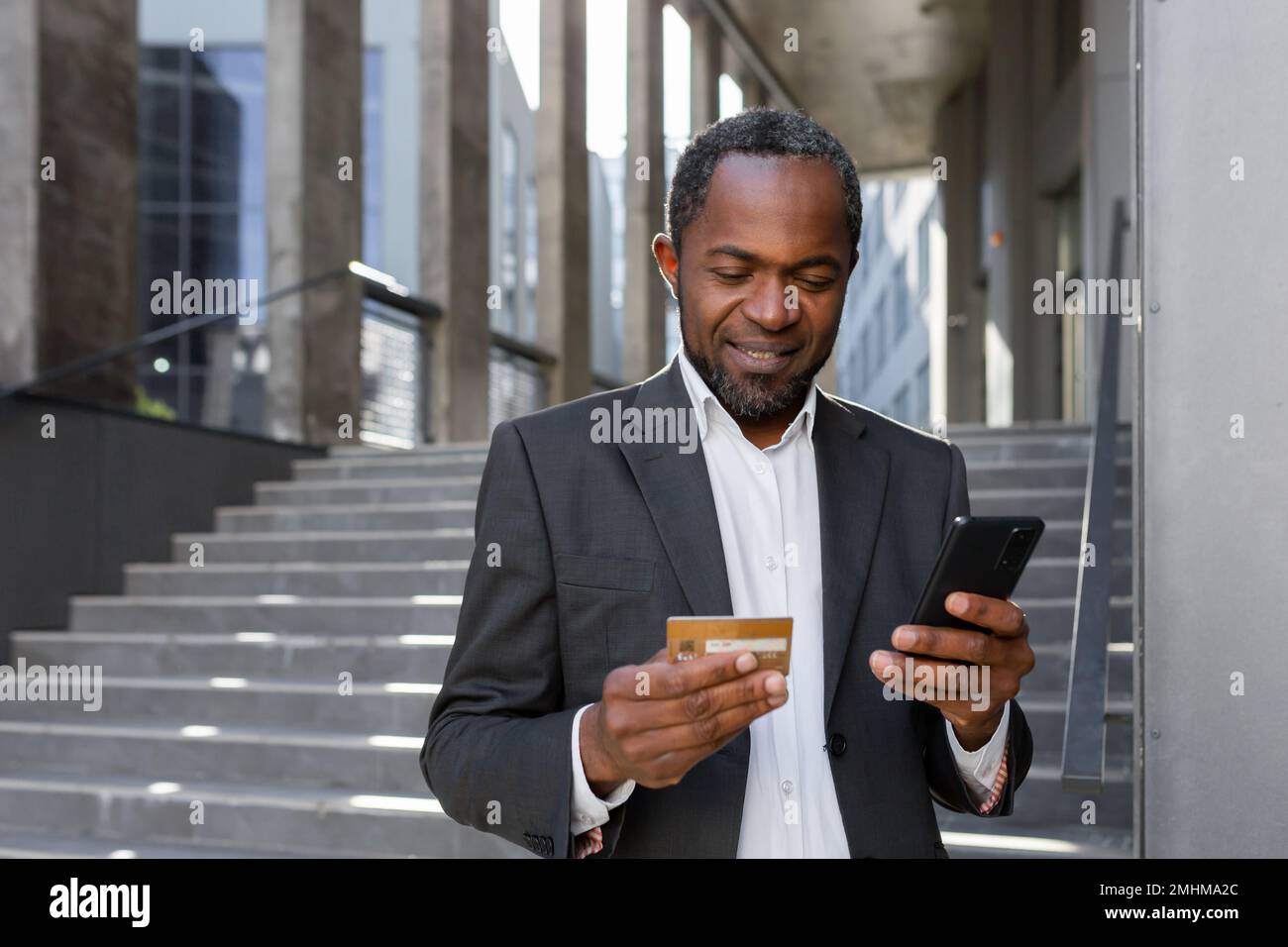 Homme afro-américain heureux et réussi en costume d'affaires à l'extérieur de bâtiment de bureau tenant la carte de crédit de banque et le téléphone, homme d'affaires shopping en ligne et de transférer de l'argent à l'aide de l'application. Banque D'Images