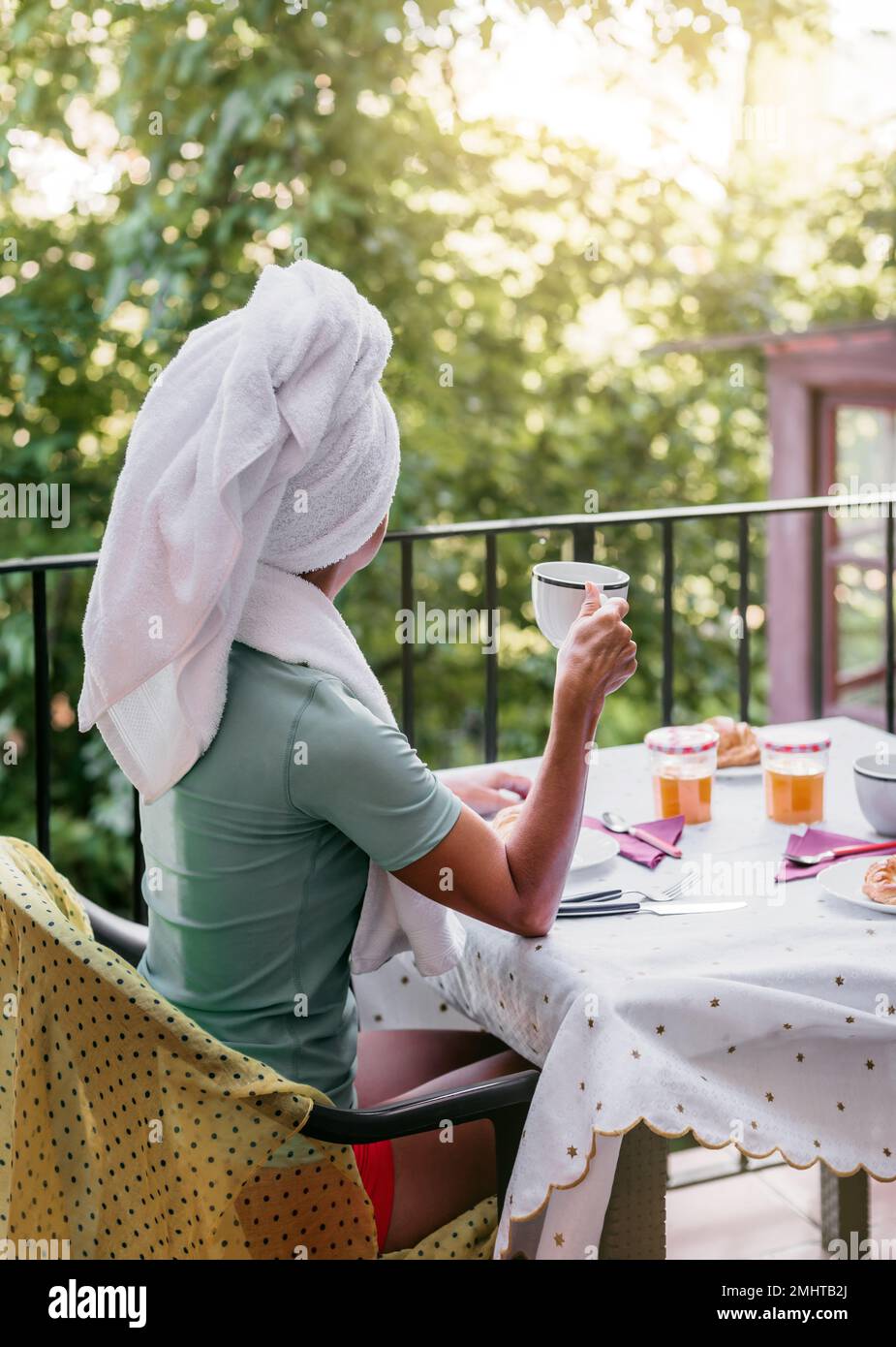 une femme de race blanche d'âge moyen prend le petit-déjeuner sur la terrasse de sa maison après une douche. elle porte une serviette sur sa tête. concept de bien-être et Banque D'Images