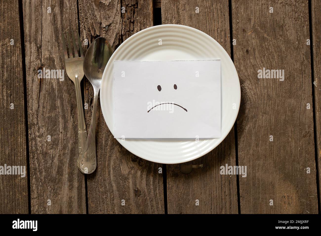 assiette vide avec un smiley triste sur papier et une fourchette avec une cuillère sur une table en bois, rien à manger Banque D'Images