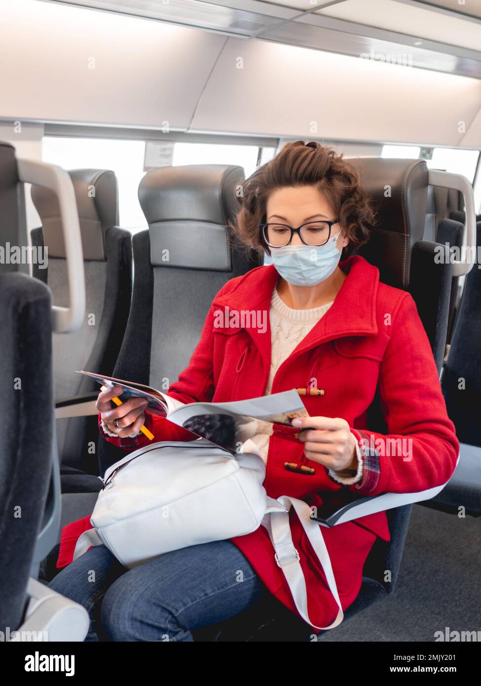 Femme en duffle-coat rouge et masque de protection lit le magazine dans le train de banlieue. Voyager en véhicule terrestre. Nouvelle normale. Banque D'Images