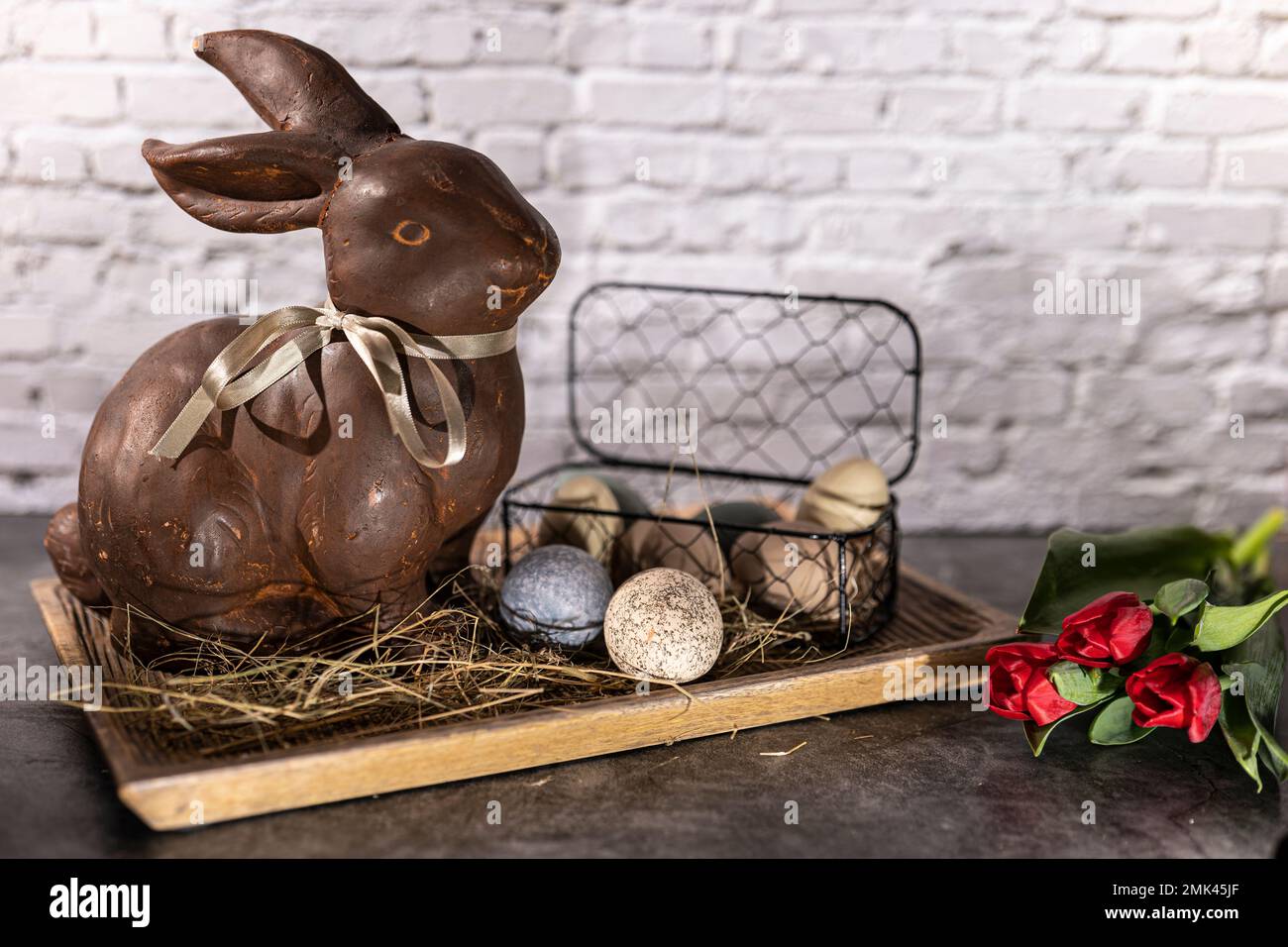 lapin de pâques en argile brune dans un bol en bois avec œufs de pâques et foin devant le mur de briques blanches avec tulipes rouges Banque D'Images