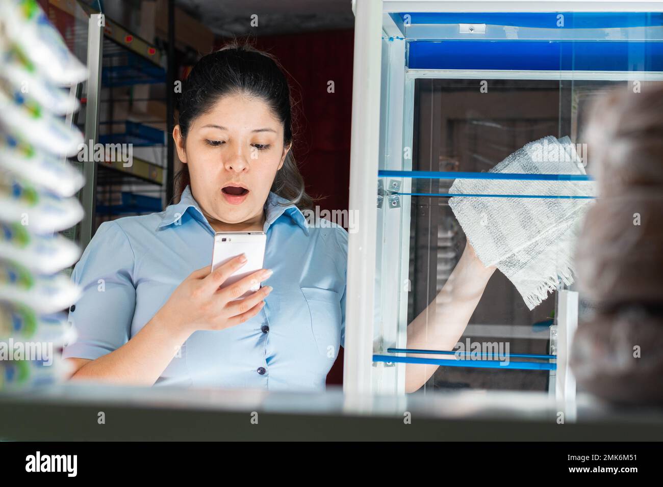 jeune latina femme, très impressionné regardant son téléphone cellulaire, tout en travaillant dans un magasin, essuyant les vitrines avec une serviette Banque D'Images