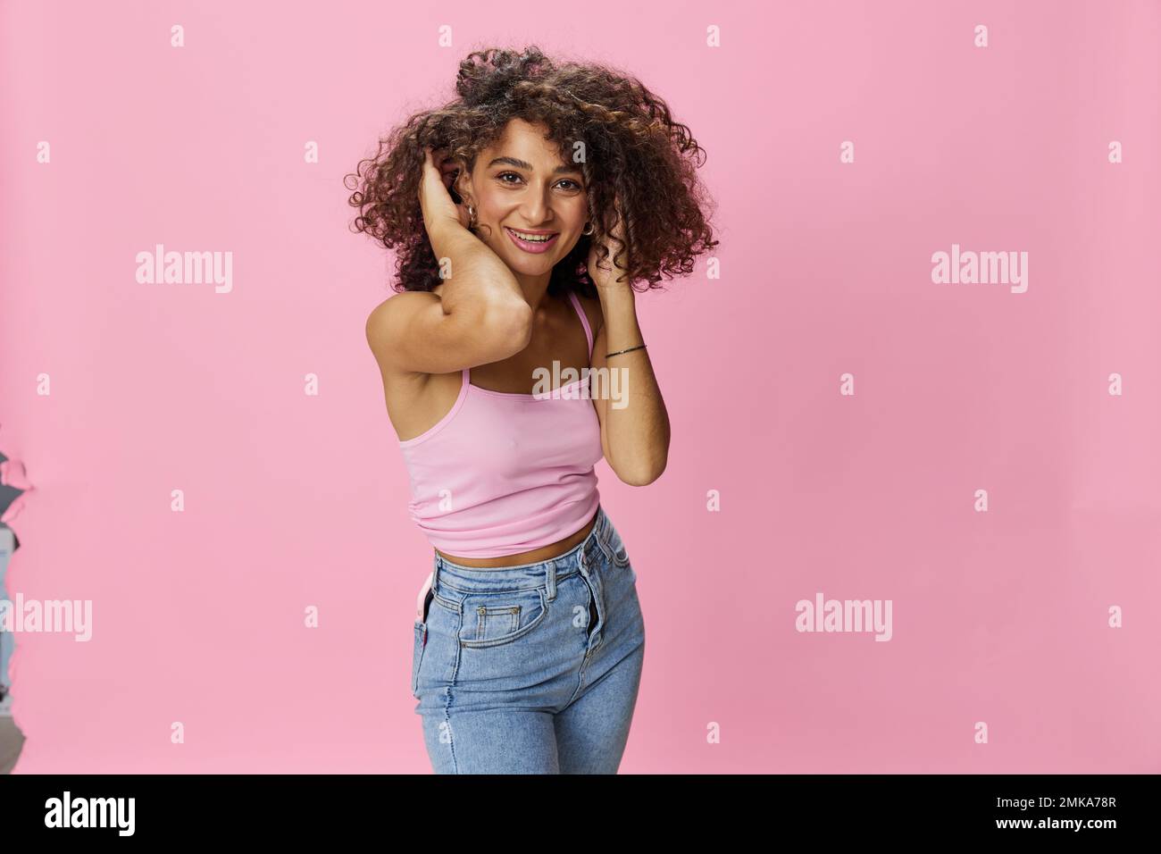 Femme avec le curly afro cheveux modèle pose sur un fond rose dans un T-shirt rose, libre mouvement et danse, regarder dans la caméra, sourire avec des dents et Banque D'Images