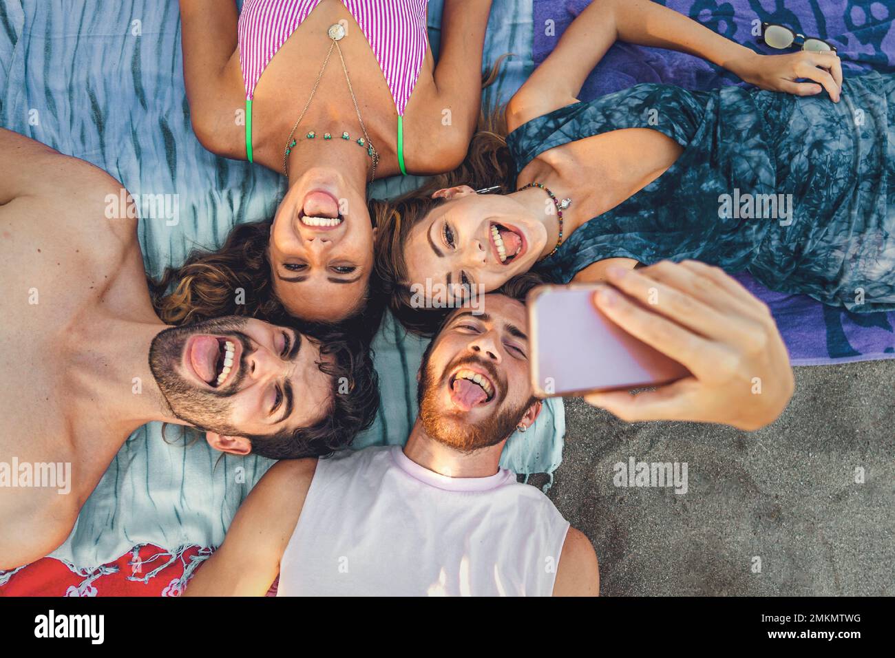 Quatre jeunes adultes qui profitent d'une journée à la plage, allongé sur des serviettes et prenant un selfie de groupe. Riant et faisant des visages stupides, ils capturent la joie et moi Banque D'Images