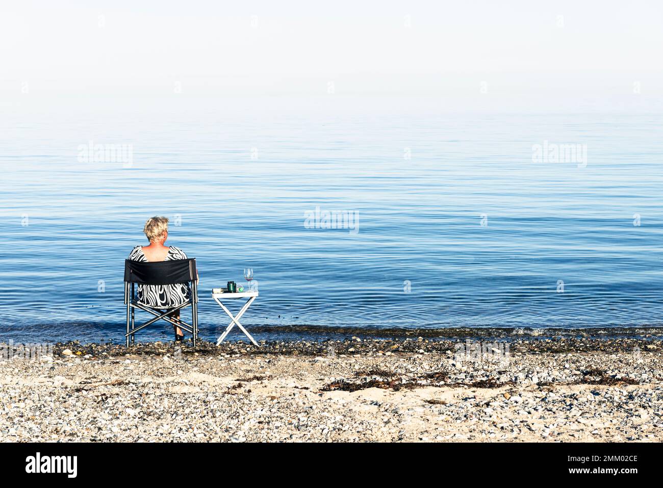 Une femme blonde est assise sur une chaise de camping sur la rive de Kattegatt au soleil et boit du vin, Djursland, Jutland, Danemark Banque D'Images