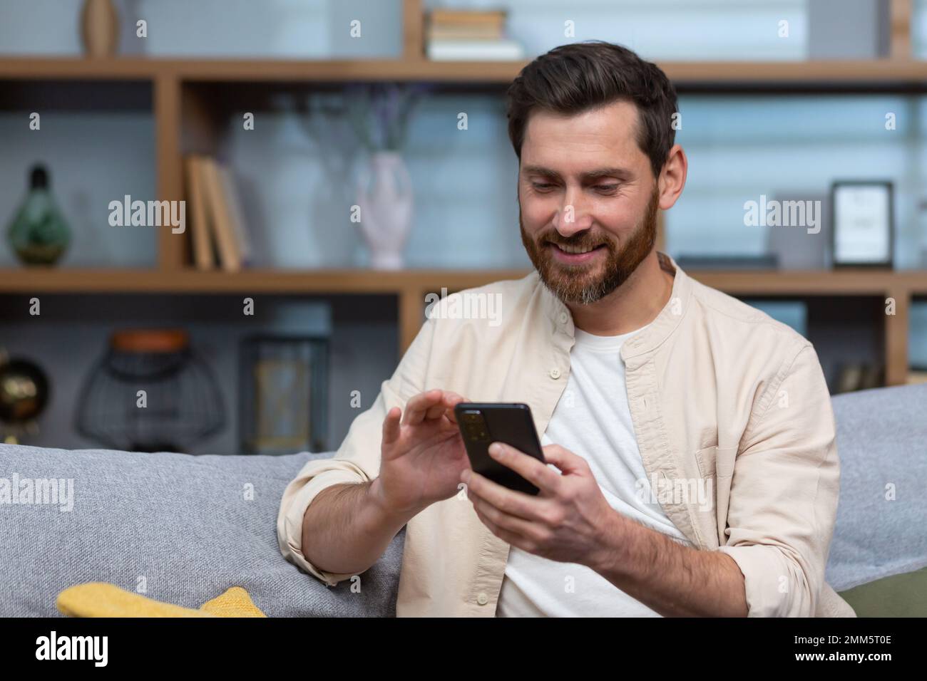 Gros plan. Un jeune homme beau dans une chemise beige est assis à la maison sur le canapé, utilise un téléphone. Banque D'Images