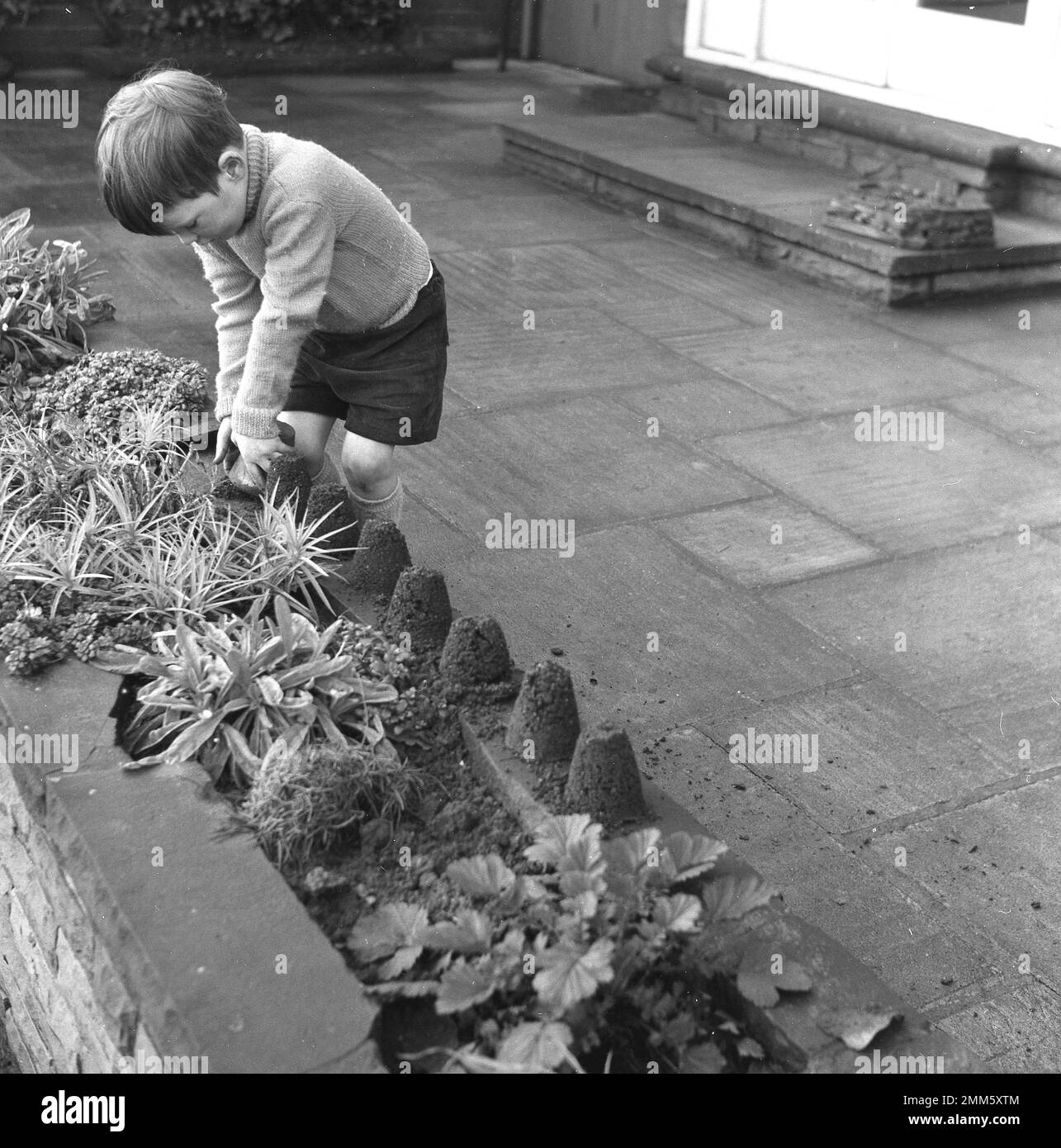 1960s, historique, un jeune garçon, en short et un pull à col roulé, jouant seul à l'extérieur sur un jardin patio, faisant de petits châteaux de sable de terre! Banque D'Images