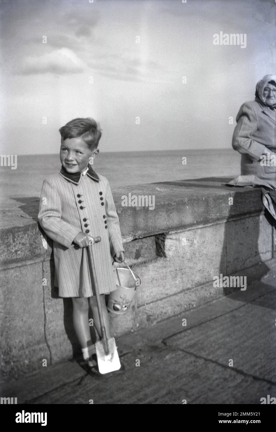 1950s, historique, penchée contre un mur de mer sur la côte, un petit garçon portant une élégante veste rayée, avec col en velours, tenant son seau et sa bêche, Bridlington, East Yorkshire, Angleterre. ROYAUME-UNI. Banque D'Images