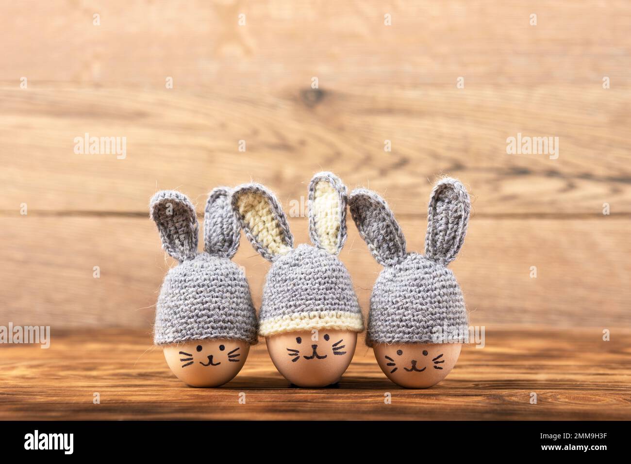 Trois œufs de Pâques aux visages amusants et chapeaux crochetés gris avec oreilles de lapin sur fond de bois. Concept joyeuses Pâques. Carte de vœux. Banque D'Images