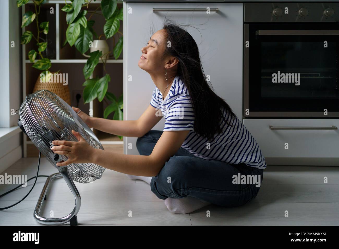 Joyeuse positive asiatique fille aime le vent froid du ventilateur électrique se trouve sur le sol dans la cuisine de la maison Banque D'Images