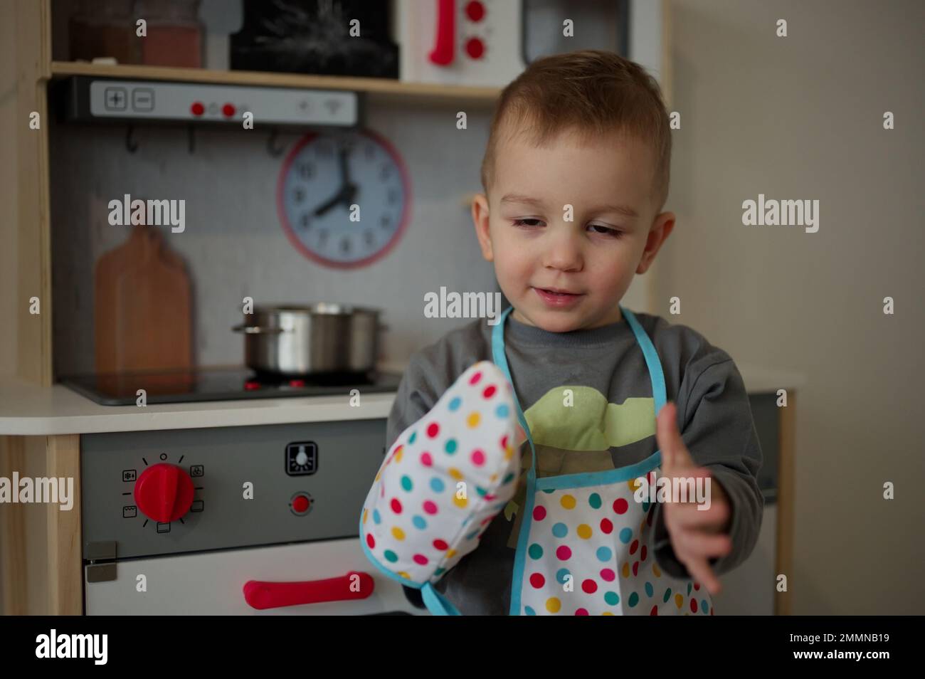 Un petit tout-petit mignon avec un tablier et un gant devant un ensemble de cuisine jouet Banque D'Images