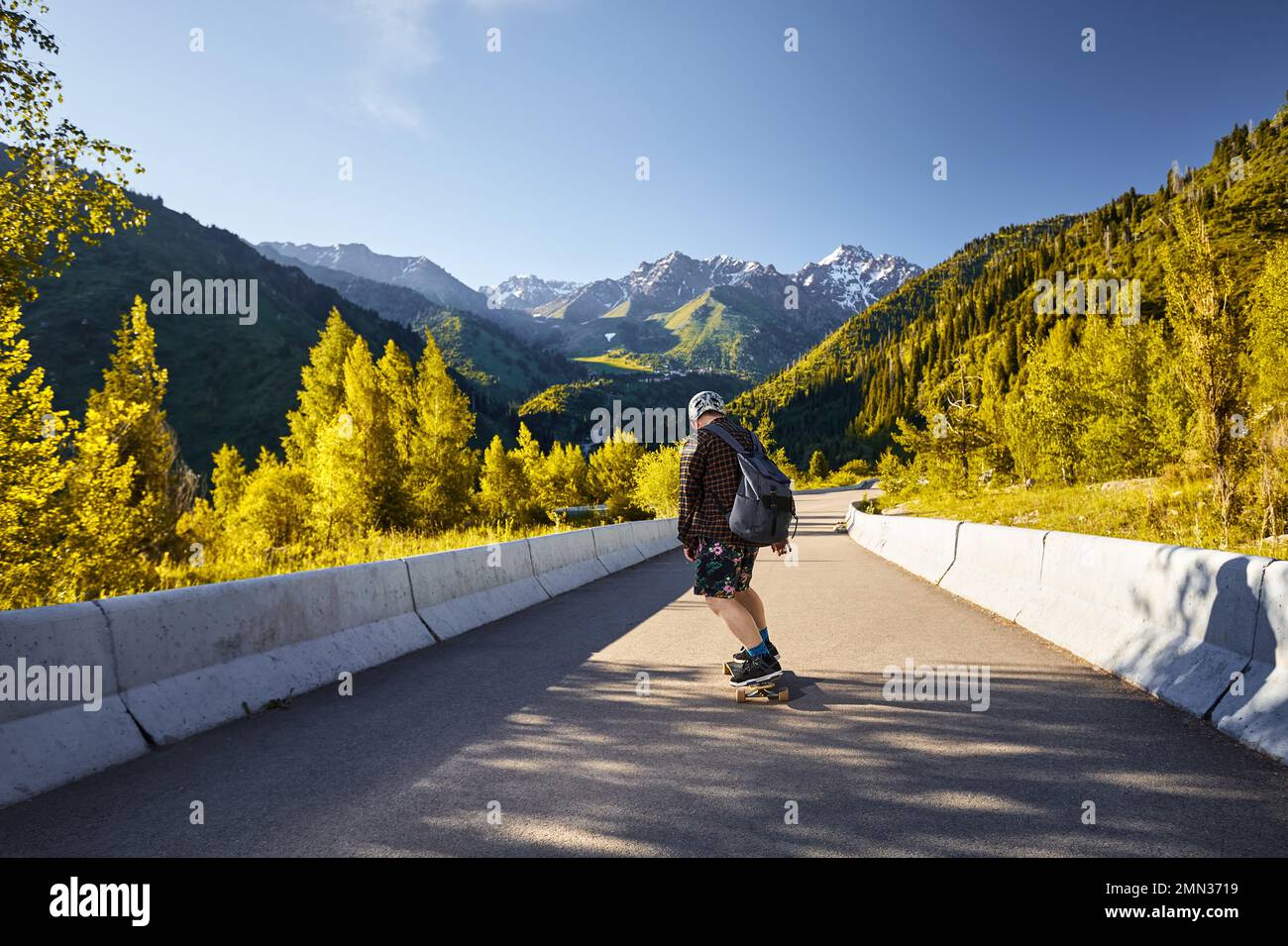 Homme Skater dans le casque et tour en chemise orange sur la route de montagne à son bord de mer près du sommet de neige au matin d'été Kazakhstan Banque D'Images