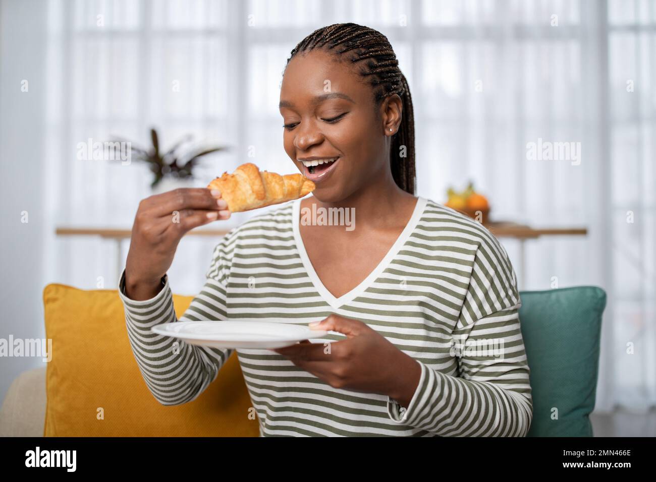 Joyeuse jeune femme noire prenant le petit déjeuner à la maison, en mangeant un croissant Banque D'Images