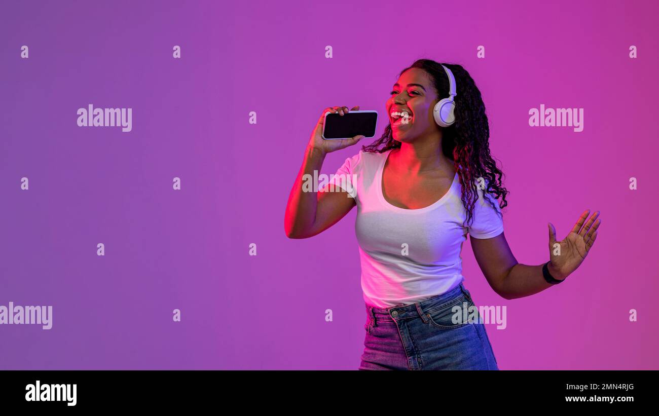 Joyeuse femme noire écoutant de la musique dans un casque sans fil et chantant avec un smartphone Banque D'Images