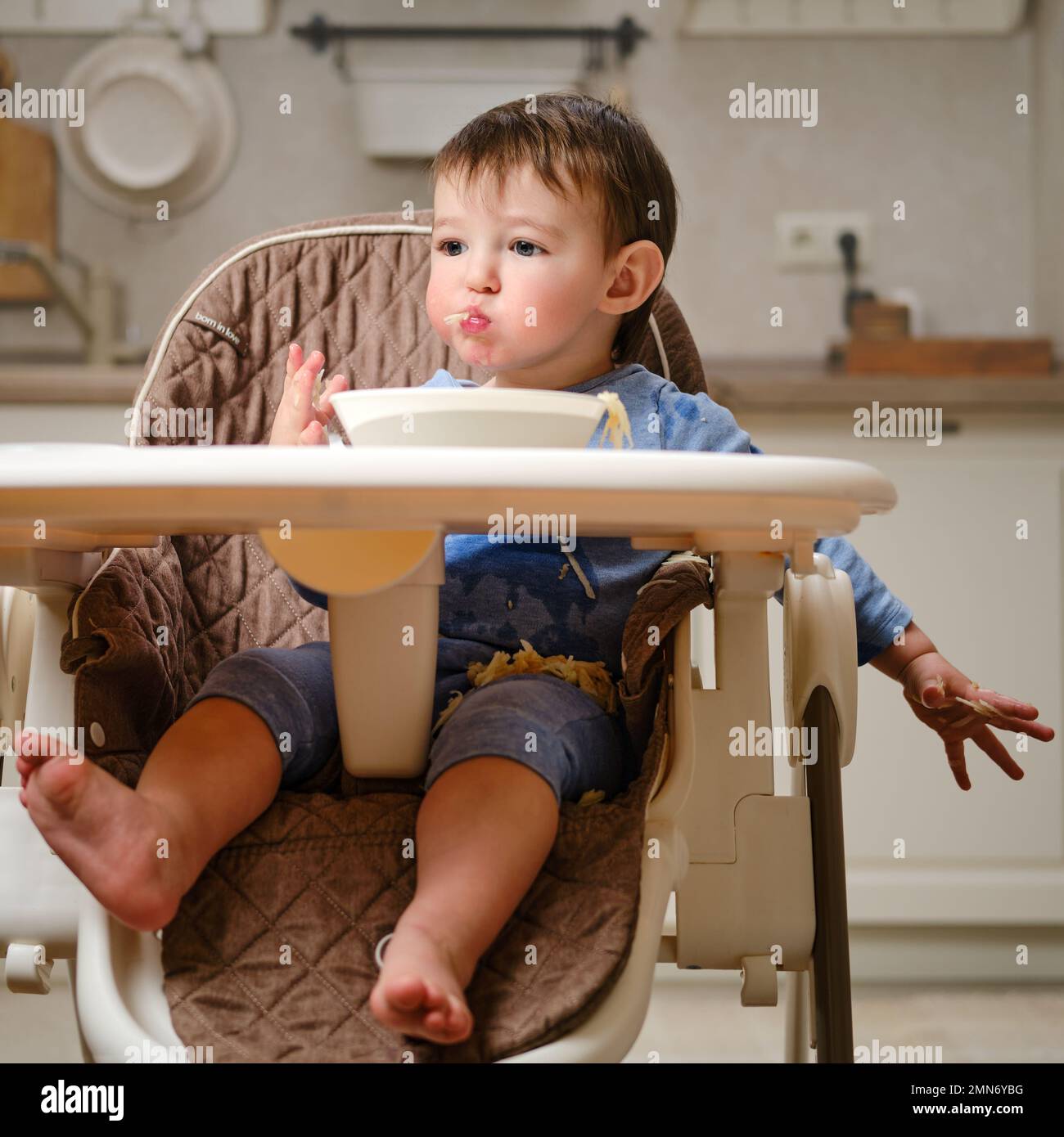 Un enfant drôle mange une pomme râpée avec sa bouche pleine tout en étant assis sur une chaise de cuisine. Bébé garçon affamé, il a de la nourriture dans sa bouche, de l'humour. Enfant âgé Banque D'Images