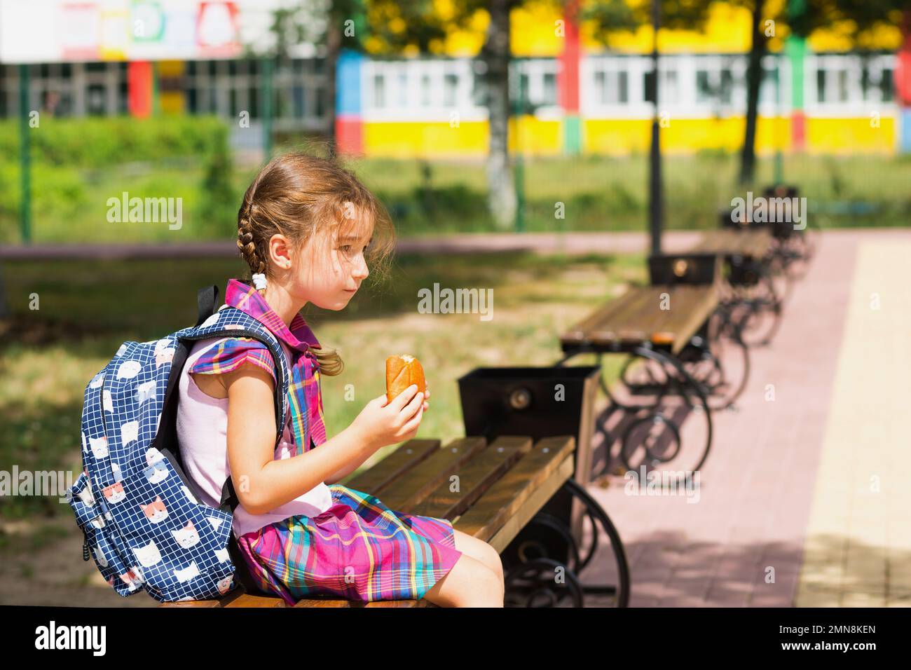 Fille avec un sac à dos assis sur un banc et manger une tarte près de l'école. Une collation rapide avec un petit pain, nourriture malsaine, déjeuner de la maison. Retour à l'école. E Banque D'Images