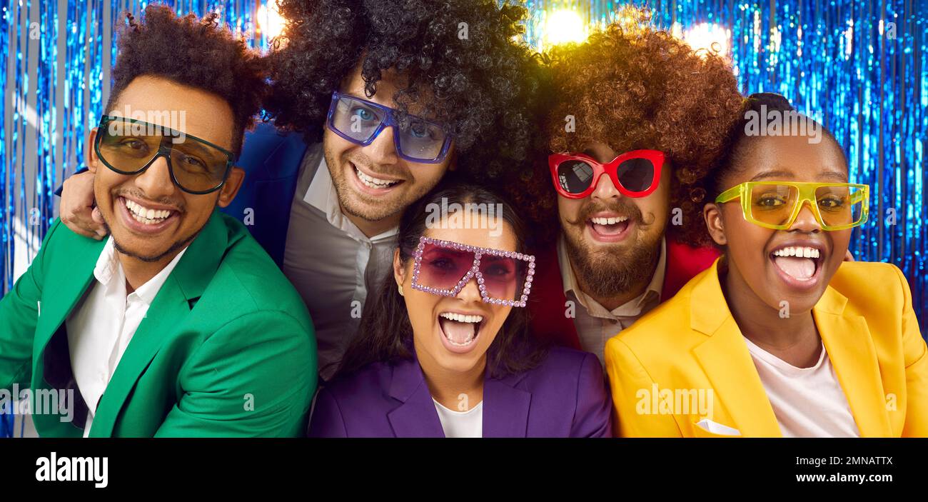 Joyeux joyeux multiracial dans des costumes colorés, des verres disco et des perruques drôles dans le stand de photo Banque D'Images