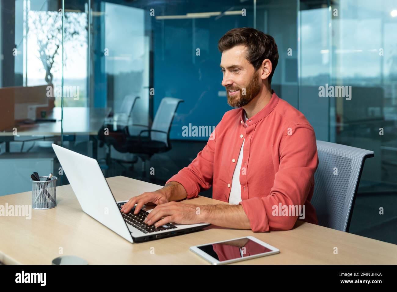 Formation à distance. Portrait d'un jeune étudiant dans une chemise rouge. Assis au bureau, étudiant en ligne sur un ordinateur portable. Écrit un diplôme, un cours, un résumé, une conférence. Banque D'Images