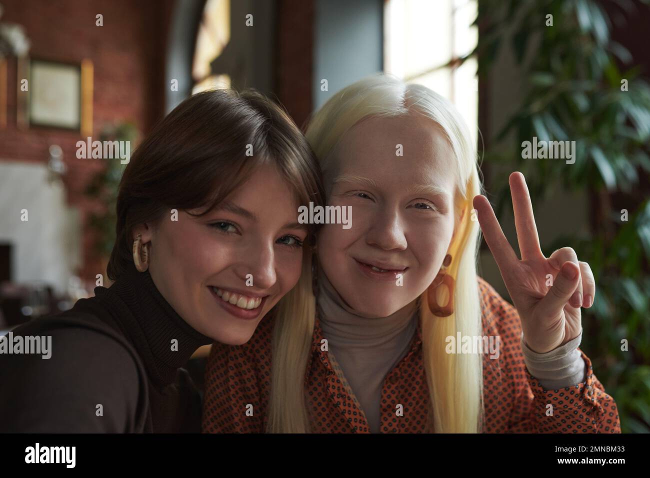 Jeune femme albino avec de longs cheveux montrant le geste de paix tout en étant assise à côté de son ami et les deux regardant l'appareil photo dans le café Banque D'Images