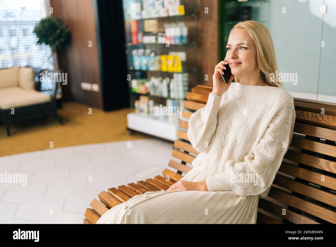 Portrait d'une jeune femme blonde élégante parlant sur un smartphone, souriant discutant des dernières nouvelles, appréciant une conversation agréable assise sur un banc en bois. Banque D'Images