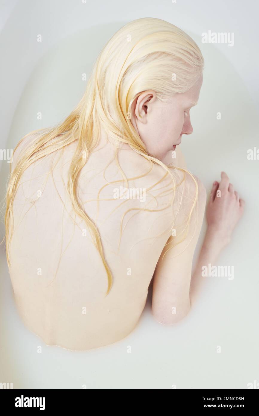 Dos de jeune femme albino fraîche avec de longs cheveux blonds mouillés prenant le bain tout en étant assis dans la baignoire avec le lait chaud et en appréciant la procédure d'hygiène Banque D'Images